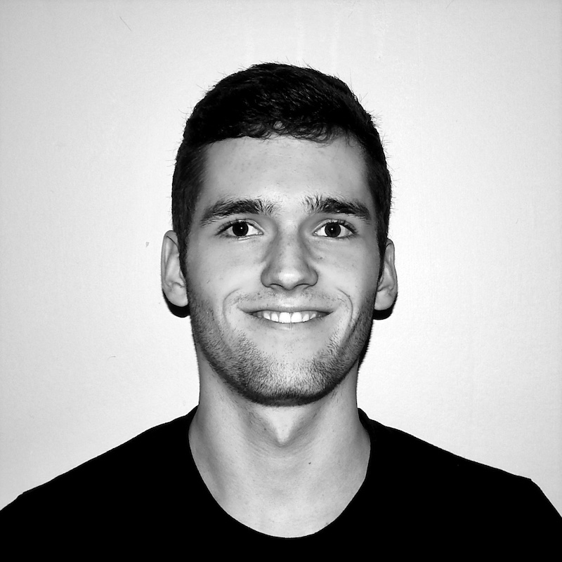 Steven Goodman, a white man with short brown hair and a slight beard is visible from the shoulders up standing in front of a solid white background and wearing a dark shirt. He is looking directly at the camera and smiling. The photo is in black and white.